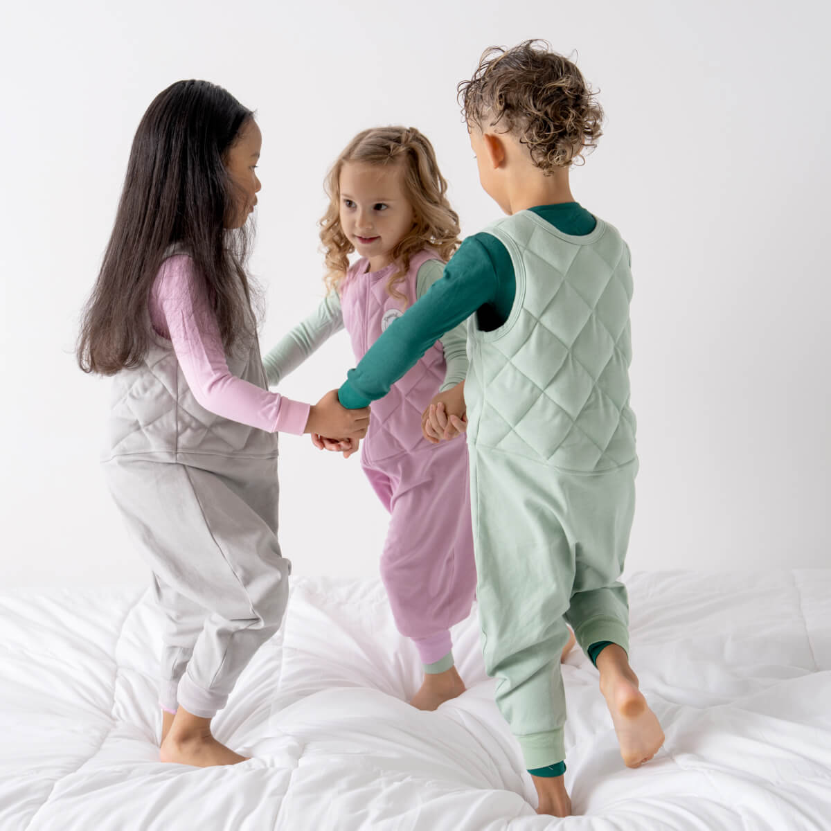 Three young children holding hands on a white bed in Dreamland Baby weighted sleep sack walkers and pajamas.